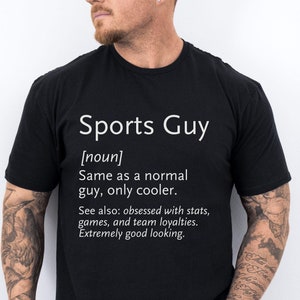 May include: Black t-shirt with white text that reads "Sports Guy [noun] Same as a normal guy, only cooler. See also: obsessed with stats, games, and team loyalties. Extremely good looking."