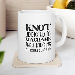 May include: White ceramic mug with the text "KNOT ADDICTED TO MACRAME JUST KIDDING I'M TOTALLY ADDICTED" in black lettering. The mug sits on a beige coaster. The background includes a wooden cutting board, a white bowl with fruit, and greenery.