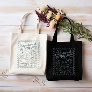 May include: Two tote bags, one cream and one black, with the text "professional yapper" in a stamp-like design. The design includes speech-related icons and the text "speech language pathologist Mrs. Smith". The bags have long handles.