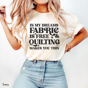 Op de afbeelding: Ivoor kleurig t-shirt met de zwarte tekst "IN MY DREAMS FABRIC IS FREE & QUILTING MAKES YOU THIN". Het shirt wordt gecombineerd met lichtblauwe jeans. De algehele stijl is casual en comfortabel.