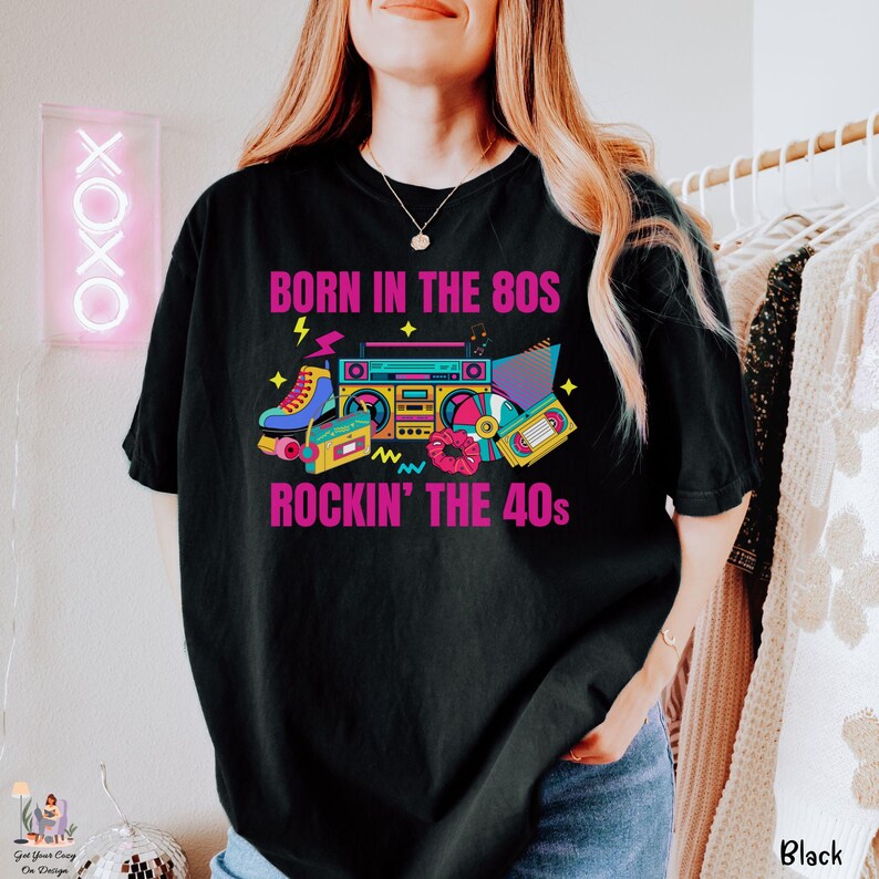 May include: Black t-shirt with a vibrant retro design. The graphic includes a boombox, roller skates, and cassette tapes, with the text "BORN IN THE 80S ROCKIN' THE 40s" in pink. A casual, comfortable style.