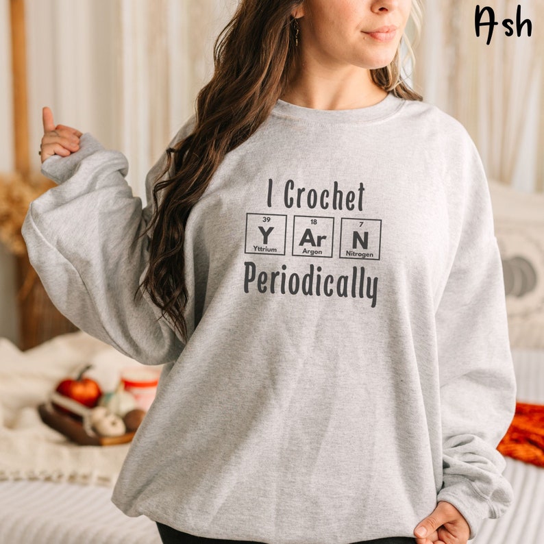 This Funny Crocheting Sweatshirt is a Gildan 18000 crewneck sweatshirt in the color Ash. The text on front says I Crochet Yarn Periodically with the word Yarn spelled out in the periodic table.