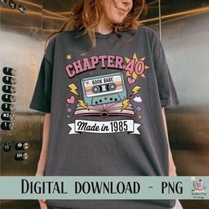 May include: Dark gray t-shirt with a colorful graphic. The graphic includes the text "CHAPTER 40" in pink, a cassette tape, and the words "Made in 1985". The shirt has a relaxed fit.