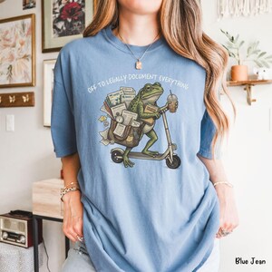 May include: Blue t-shirt with the text "OFF TO LEGALLY DOCUMENT EVERYTHING." The graphic shows a green frog riding a scooter, carrying documents and a coffee cup. The shirt is a light blue color.