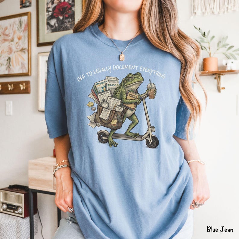 May include: Blue t-shirt with the text "OFF TO LEGALLY DOCUMENT EVERYTHING." The graphic shows a green frog riding a scooter, carrying documents and a coffee cup. The shirt is a light blue color.