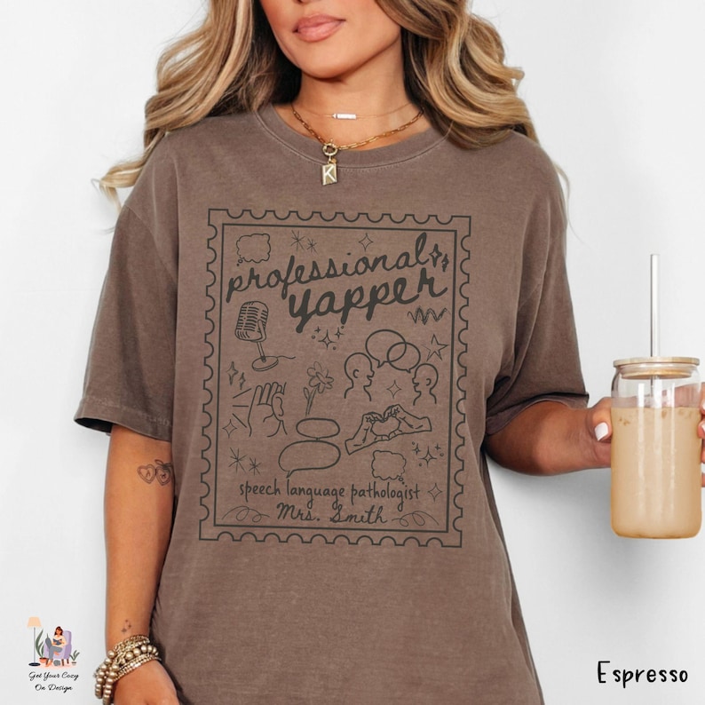 以下が含まれることがあります： エスプレッソ色のTシャツ。切手デザインの中に「professional yapper」と書かれたグラフィックがプリントされています。グラフィックには、マイク、吹き出し、その他の落書きが含まれています。「speech language pathologist」と「Mrs. Smith」の文字も含まれています。