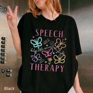 Puede incluir: Camiseta negra con las palabras "SPEECH THERAPY" en rosa y gr&aacute;ficos de mariposas coloridas. Tambi&eacute;n se incluye el texto "helps little voices take flight". La camiseta es de estilo casual.