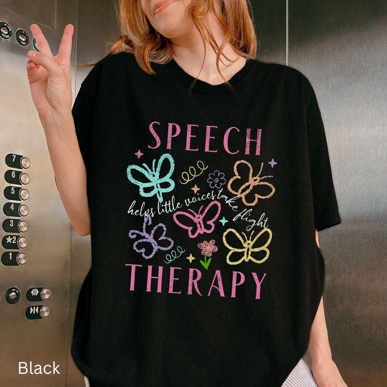 Puede incluir: Camiseta negra con las palabras "SPEECH THERAPY" en rosa y gr&aacute;ficos de mariposas coloridas. Tambi&eacute;n se incluye el texto "helps little voices take flight". La camiseta es de estilo casual.
