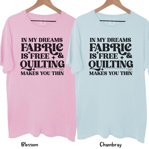 Op de afbeelding: Twee T-shirts met korte mouwen, &eacute;&eacute;n lichtroze en &eacute;&eacute;n lichtblauw, hangen aan houten hangers. Elk shirt heeft de tekst "IN MY DREAMS FABRIC IS FREE & QUILTING MAKES YOU THIN" in het zwart. Het roze shirt is gelabeld "Blossom" en het blauwe shirt "Chambray".