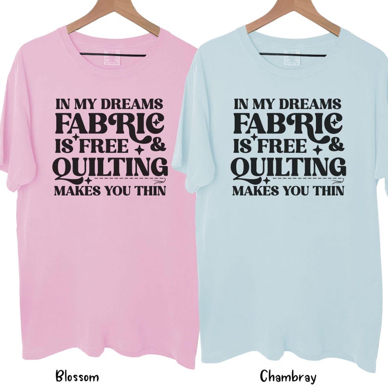 Op de afbeelding: Twee T-shirts met korte mouwen, &eacute;&eacute;n lichtroze en &eacute;&eacute;n lichtblauw, hangen aan houten hangers. Elk shirt heeft de tekst "IN MY DREAMS FABRIC IS FREE & QUILTING MAKES YOU THIN" in het zwart. Het roze shirt is gelabeld "Blossom" en het blauwe shirt "Chambray".