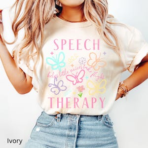 Puede incluir: Camiseta de color marfil con las palabras "SPEECH THERAPY" en rosa, junto con gr&aacute;ficos coloridos de mariposas y flores. La camiseta tambi&eacute;n presenta la frase "helps little voices take flight".