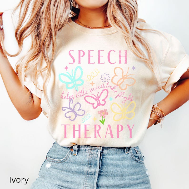 Puede incluir: Camiseta de color marfil con las palabras "SPEECH THERAPY" en rosa, junto con gr&aacute;ficos coloridos de mariposas y flores. La camiseta tambi&eacute;n presenta la frase "helps little voices take flight".