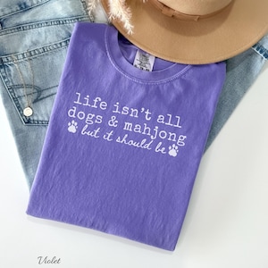 May include: A violet short-sleeved t-shirt featuring the white printed text "life isn't all dogs & mahjong but it should be" accompanied by two small paw print icons. The shirt is layered with light blue denim jeans and a tan wide-brimmed hat.