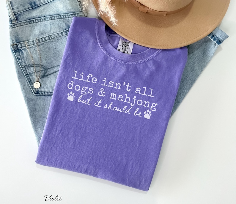 May include: A violet short-sleeved t-shirt featuring the white printed text "life isn't all dogs & mahjong but it should be" accompanied by two small paw print icons. The shirt is layered with light blue denim jeans and a tan wide-brimmed hat.