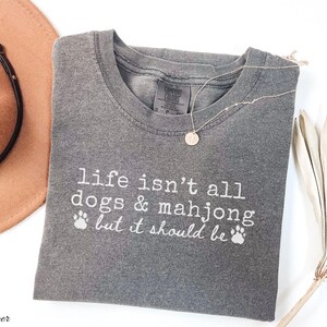 May include: A charcoal gray short-sleeve t-shirt with white text that reads, "life isn't all dogs & mahjong but it should be," accompanied by two small paw print icons. The shirt features a crew neckline and a visible brand label at the inner collar.