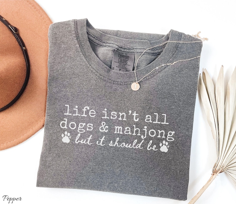 May include: A charcoal gray short-sleeve t-shirt with white text that reads, "life isn't all dogs & mahjong but it should be," accompanied by two small paw print icons. The shirt features a crew neckline and a visible brand label at the inner collar.
