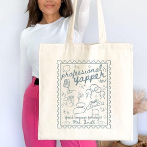 May include: Cream-colored tote bag with a blue stamp-like design. The design includes the words "professional yapper" and "speech language pathologist Mrs. Smith" along with illustrations of a microphone, speech bubbles, and other symbols.