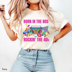 May include: Ivory t-shirt with a vibrant graphic. The design includes a boombox, roller skates, cassette tapes, and the text "BORN IN THE 80S ROCKIN' THE 40S" in pink and yellow. The shirt is worn with blue denim jeans.