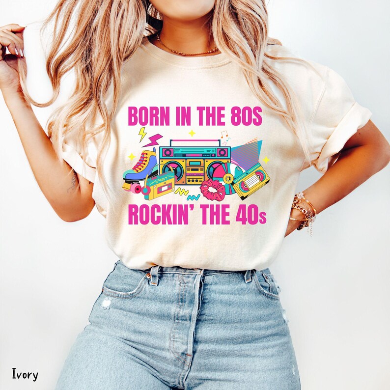 May include: Ivory t-shirt with a vibrant graphic. The design includes a boombox, roller skates, cassette tapes, and the text "BORN IN THE 80S ROCKIN' THE 40S" in pink and yellow. The shirt is worn with blue denim jeans.