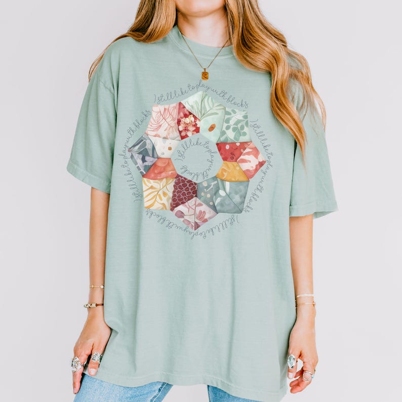 May include: A light green t-shirt with a circular design featuring a patchwork quilt pattern in shades of red, orange, yellow, green, blue, and brown. The design is surrounded by the text "I'll like to play with blocks".