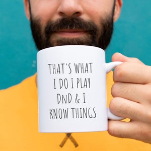 May include: White ceramic mug with black text that reads "That's what I do I play DND & I know things".