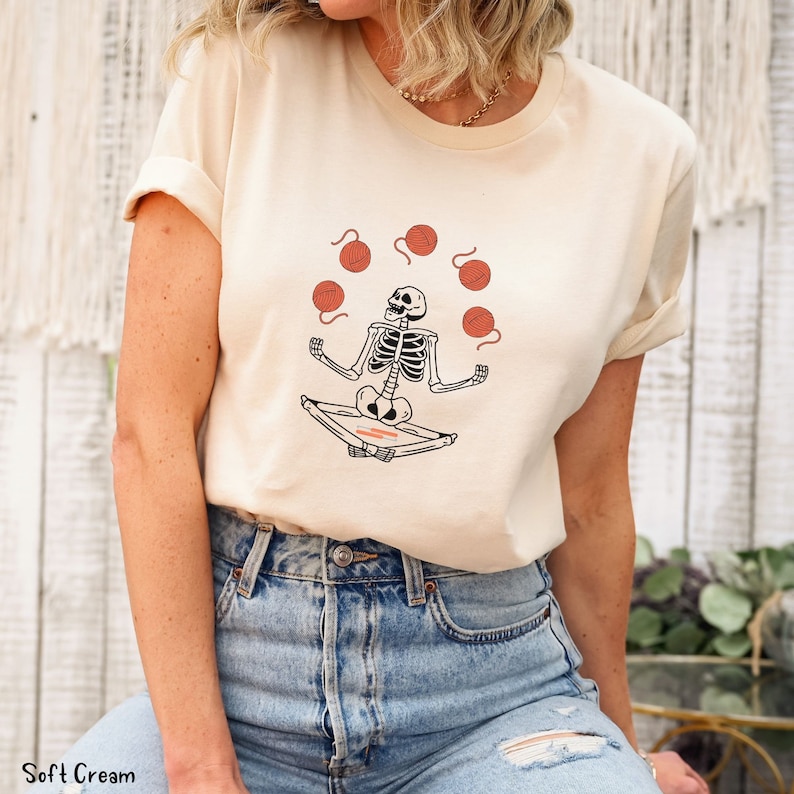May include: A cream colored t-shirt with a skeleton meditating and juggling balls of yarn. The yarn is red and the skeleton is black.