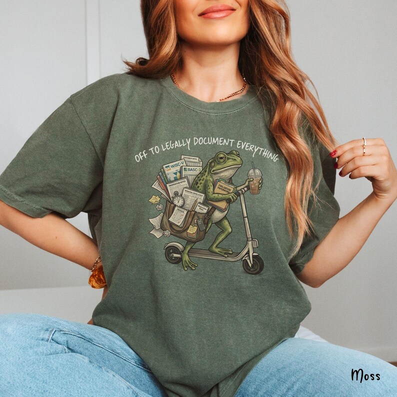 May include: Moss green t-shirt with the phrase "OFF TO LEGALLY DOCUMENT EVERYTHING." The graphic features a frog riding a scooter, carrying documents and a coffee cup. The shirt is a relaxed fit.