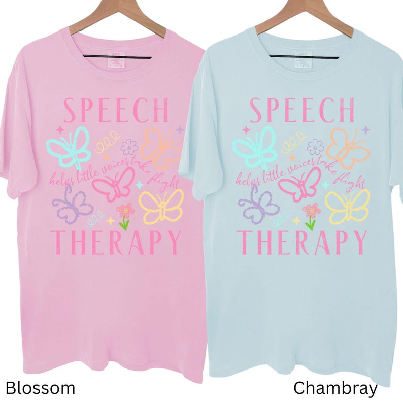 Puede incluir: Dos camisetas de manga corta, una rosa flor y otra azul chambray, cuelgan de perchas de madera. Cada camiseta presenta las palabras "SPEECH THERAPY" con gr&aacute;ficos de mariposas y flores. Tambi&eacute;n se incluye el texto "helps little voices take flight".