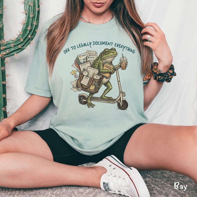 May include: A light blue t-shirt with the text "OFF TO LEGALLY DOCUMENT EVERYTHING". The shirt features a cartoon frog riding a scooter, carrying a bag of documents and a coffee cup. The frog is green, and the scooter is gray.