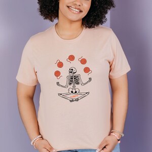 May include: A pink t-shirt with a graphic of a skeleton meditating and juggling balls of yarn.