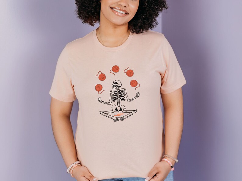 May include: A pink t-shirt with a graphic of a skeleton meditating and juggling balls of yarn.