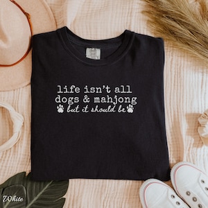 May include: A black crew neck t-shirt featuring white text that reads, life isn't all dogs & mahjong but it should be, accompanied by two small white paw print icons. The shirt has a standard fit with short sleeves and a ribbed neckline.
