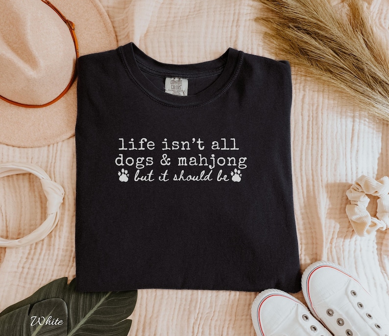 May include: A black crew neck t-shirt featuring white text that reads, life isn't all dogs & mahjong but it should be, accompanied by two small white paw print icons. The shirt has a standard fit with short sleeves and a ribbed neckline.