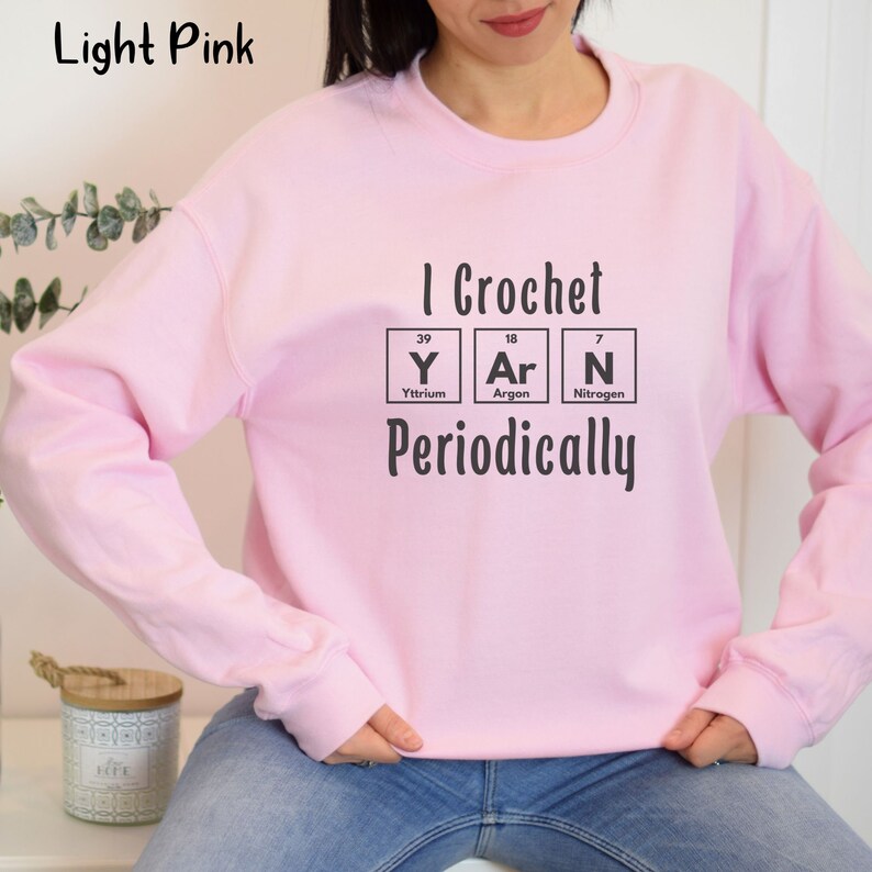 This Funny Crocheting Sweatshirt is a Gildan 18000 crewneck sweatshirt in the color Light Pink. The text on front says I Crochet Yarn Periodically with the word Yarn spelled out in the periodic table.