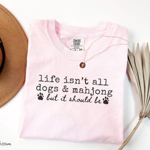May include: A light pink t-shirt featuring the black printed text "life isn't all dogs & mahjong but it should be" with two small paw print icons. The shirt has a Comfort Colors brand label at the inner neckline and a crew neck collar design.