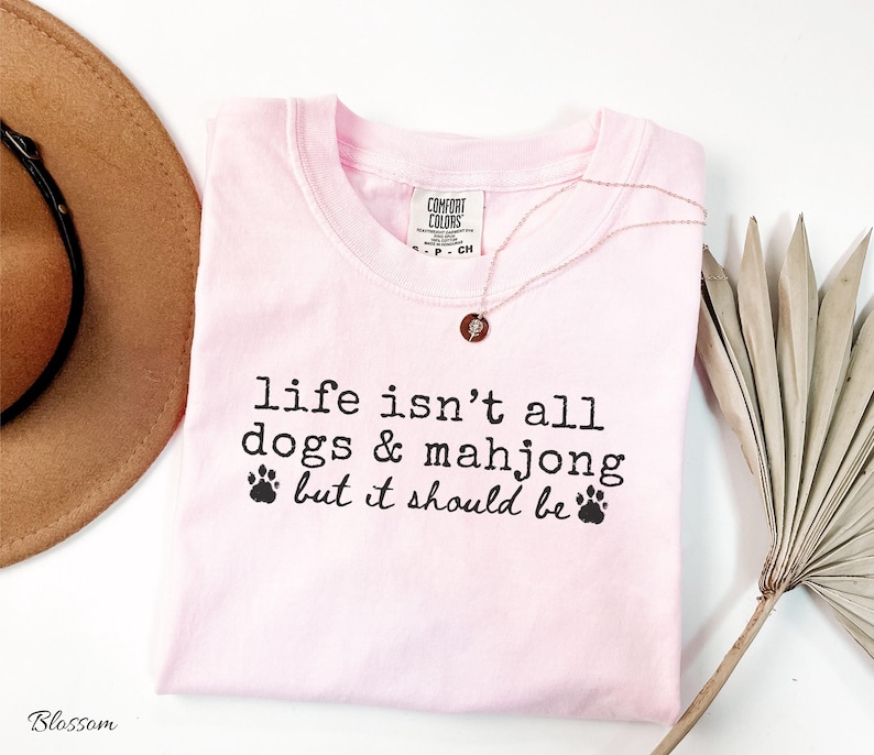 May include: A light pink t-shirt featuring the black printed text "life isn't all dogs & mahjong but it should be" with two small paw print icons. The shirt has a Comfort Colors brand label at the inner neckline and a crew neck collar design.