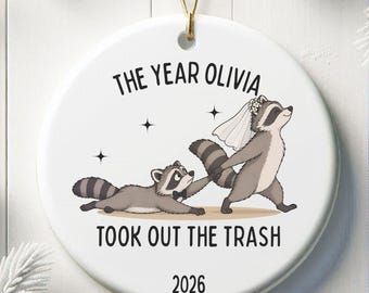 May include: A round white ceramic ornament featuring a cartoon illustration of two raccoons, one wearing a wedding veil and the other a bowtie. The text reads THE YEAR OLIVIA TOOK OUT THE TRASH and the year 2026. It hangs from a gold string.