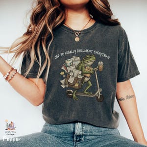 May include: Dark gray t-shirt with the text "OFF TO LEGALLY DOCUMENT EVERYTHING." The graphic shows a green frog riding a scooter, carrying documents and a coffee cup. The frog has glasses and a book. The shirt has a vintage, worn look.