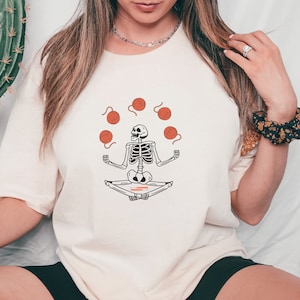 May include: A white t-shirt with a graphic of a skeleton sitting in a meditative pose, juggling balls of yarn. The yarn is orange.
