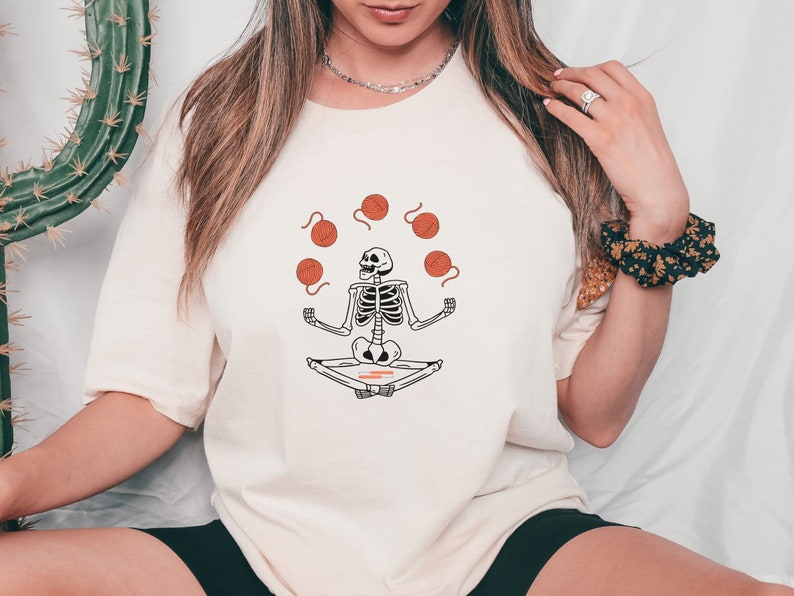 May include: A white t-shirt with a graphic of a skeleton sitting in a meditative pose, juggling balls of yarn. The yarn is orange.