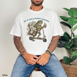 May include: White t-shirt with the text "OFF TO LEGALLY DOCUMENT EVERYTHING." The shirt features a cartoon frog riding a scooter, carrying documents and a coffee cup. The frog is green, and the scooter is gray.