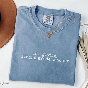 May include: A blue jean Comfort Colors t-shirt with the text "it's giving second grade teacher" in white. A gold necklace with a pendant rests on the shirt. A brown hat and dried plant are in the background.