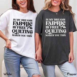 Op de afbeelding: Twee t-shirts, &eacute;&eacute;n witte en &eacute;&eacute;n donkergrijze, met de tekst "IN MY DREAMS FABRIC IS FREE & QUILTING MAKES YOU THIN" in een zwarte, vette letter. De shirts zijn casual en hebben korte mouwen.