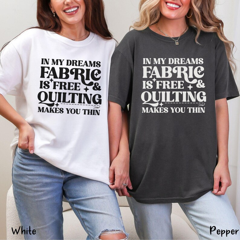 Op de afbeelding: Twee t-shirts, &eacute;&eacute;n witte en &eacute;&eacute;n donkergrijze, met de tekst "IN MY DREAMS FABRIC IS FREE & QUILTING MAKES YOU THIN" in een zwarte, vette letter. De shirts zijn casual en hebben korte mouwen.