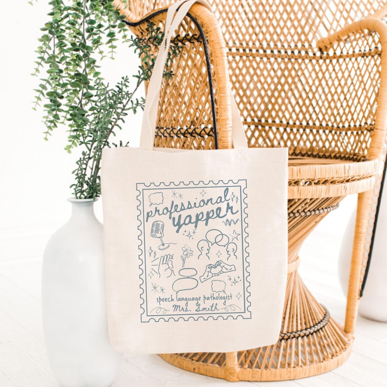 May include: A cream-colored tote bag with a blue stamp-like design that reads "professional yapper" with speech therapy-related illustrations. The bag has long handles and is made of a sturdy fabric.