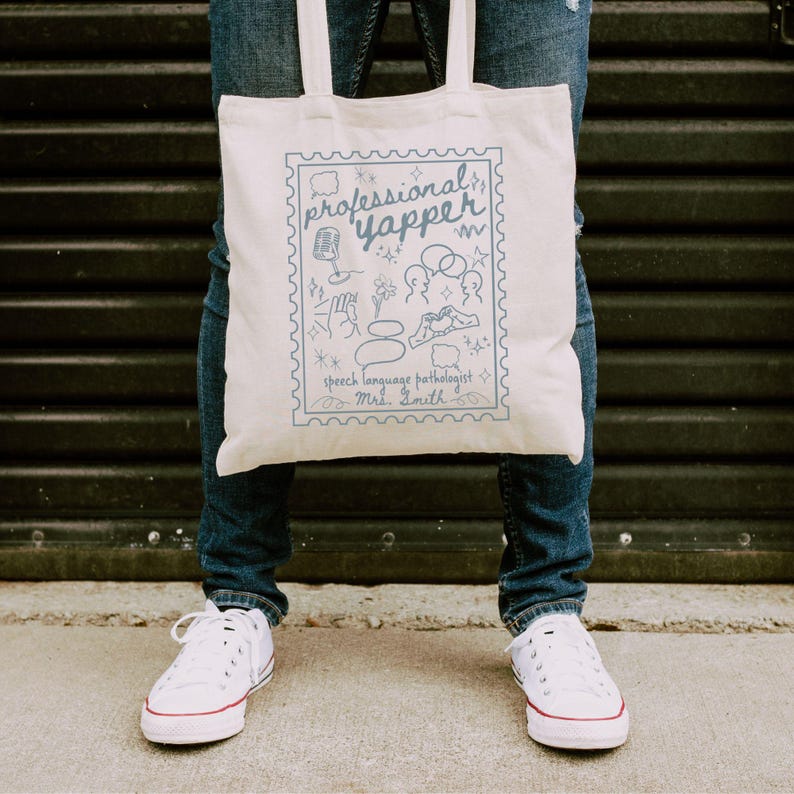 May include: A cream-colored tote bag with a blue illustration and text that reads "professional yapper, speech language pathologist, Mrs. Smith." The design includes a microphone, speech bubbles, and other related doodles. The bag has two white handles.