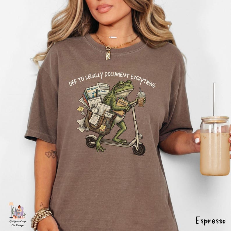 May include: Espresso-colored t-shirt featuring a whimsical illustration of a frog riding a scooter, carrying documents and a coffee. The text "OFF TO LEGALLY DOCUMENT EVERYTHING" is printed above the frog. The shirt is a comfortable, casual style.