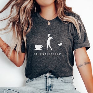 Op de afbeelding: Een donkergrijs T-shirt met een witte afbeelding van een vrouw die golf speelt, een kopje koffie en een glas wijn. De tekst "THE PLAN FOR TODAY" staat onder de afbeelding.