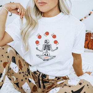 May include: White t-shirt with a graphic of a skeleton meditating and juggling balls of yarn. The skeleton is wearing a red headband.