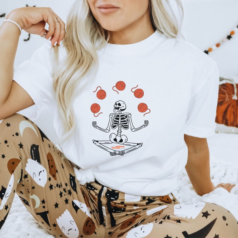 May include: White t-shirt with a graphic of a skeleton meditating and juggling balls of yarn. The skeleton is wearing a red headband.